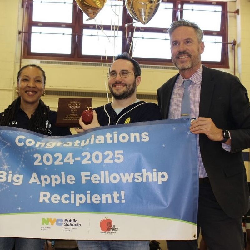 Dan holding a plaque and a red apple behind a banner that reads Congratulations, 2024-2025 Big Apple Fellowship Recipient, with two colleagues.