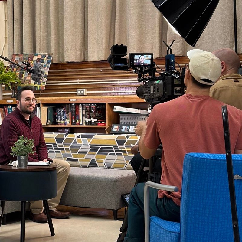 Dan seated in a school library while a film crew records an Illustrative Mathematics interview.