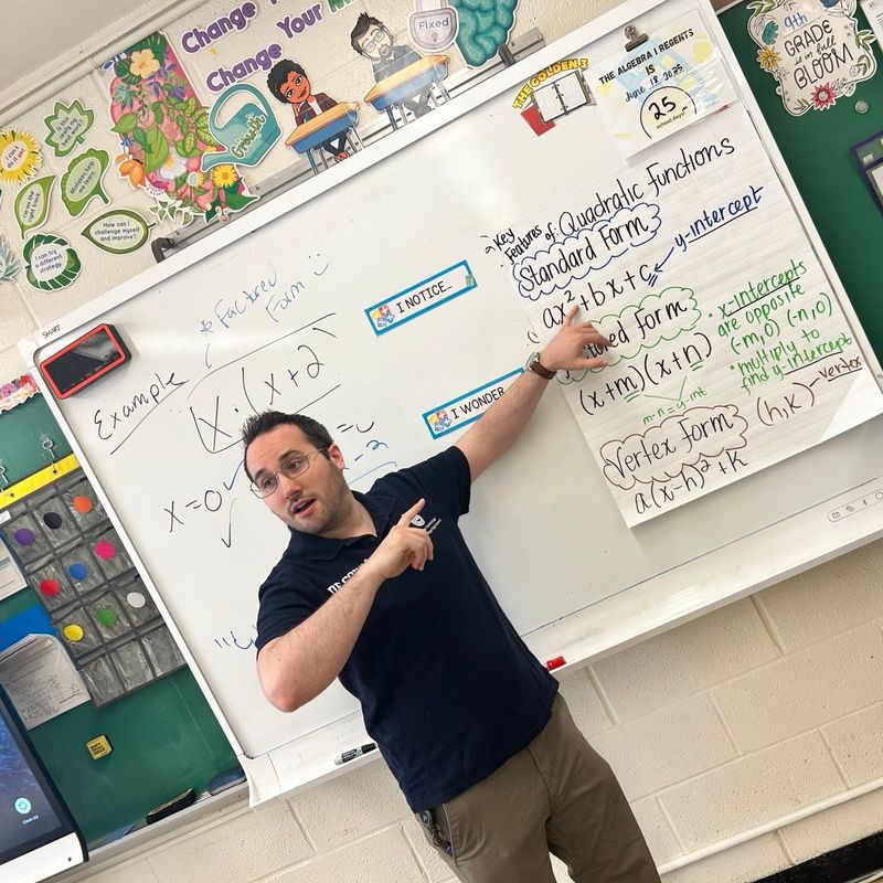 Dan at the whiteboard pointing to an anchor chart on standard, factored and vertex form.